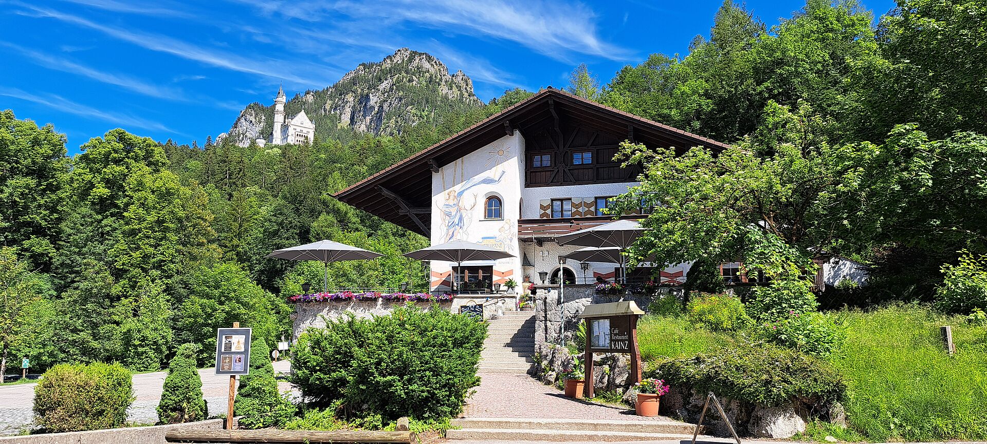 Summer photo of the Café Kainz restaurant, surrounded by green trees, with Neuschwanstein Castle in the background.