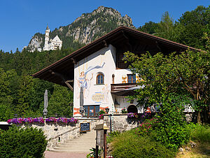 Summer view of the Café Kainz restaurant, surrounded by green trees, with Neuschwanstein Castle in the background.