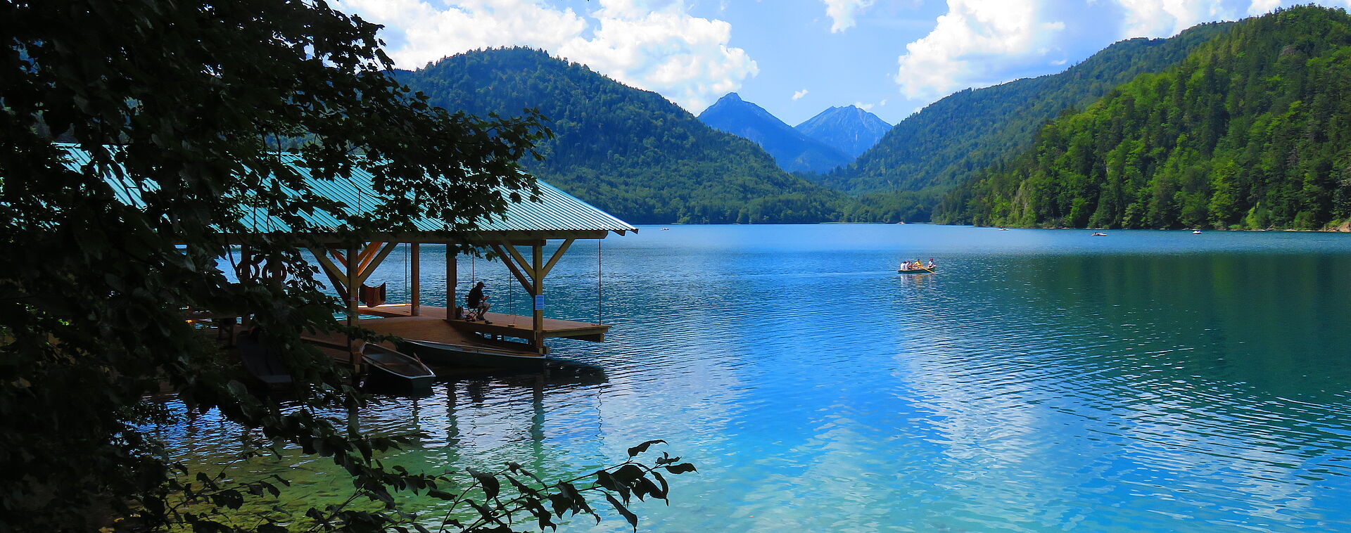 Summer shot of the boathouse on Alpsee lake with crystal clear, turquoise blue water, surrounded by green deciduous forest.