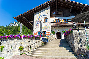 Summer photo of the Café Kainz restaurant, surrounded by green trees, with Neuschwanstein Castle in the background.