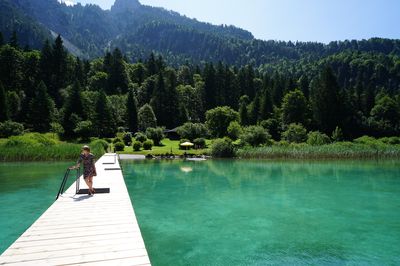 A woman stands on the wooden pier over Lake Alpsee. In the background are the sunbathing lawn with swimmers and the kiosk, surrounded by trees and shrubs.