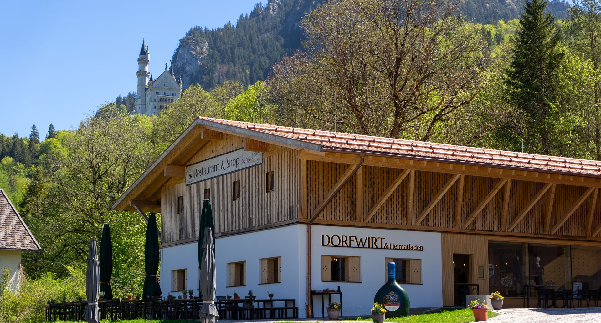 Abbildung vom Rest. Dorfwirt und Schloss Neuschwanstein im Hintergrund. Bestuhlte Terrasse mit Sonnenschirme.